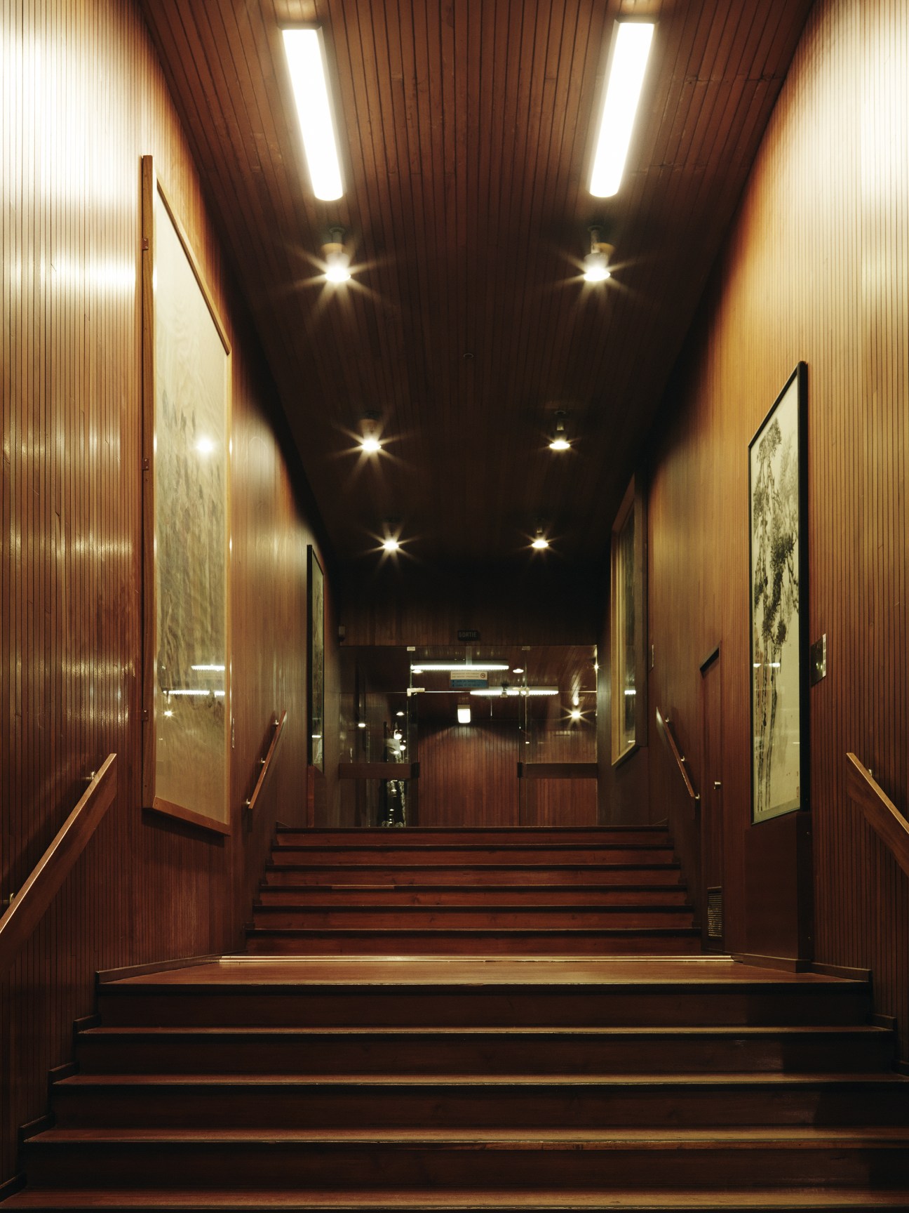 Modernist corridor with wood-paneled walls, recessed lighting, and stairs leading to a conference hall.