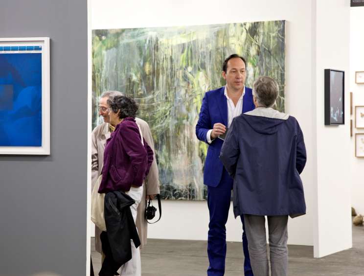 Gallery visitors and a man in a blue suit viewing artwork at an art fair.