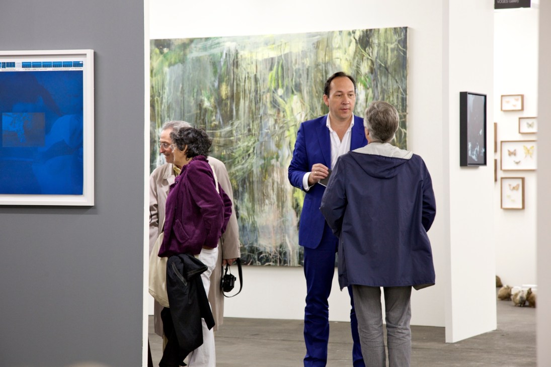 Gallery visitors and a man in a blue suit viewing artwork at an art fair.