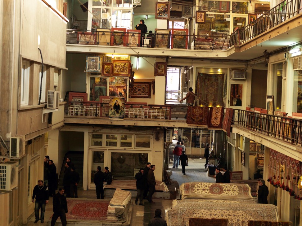 Interior view of Tehran's carpet bazaar with multiple levels displaying ornate rugs and shoppers browsing.