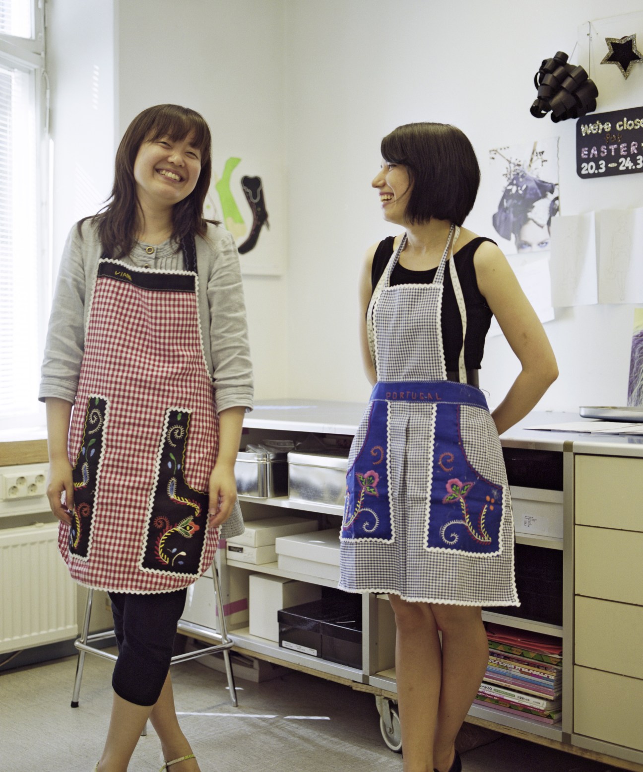Two young women wearing decorative aprons smile in a kitchen workspace.