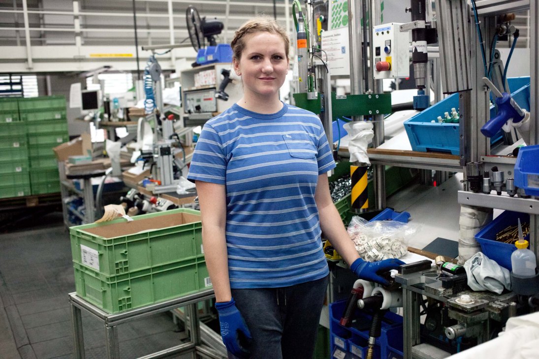 Woman in blue striped shirt standing in Hansgrohe manufacturing facility with machinery and equipment.