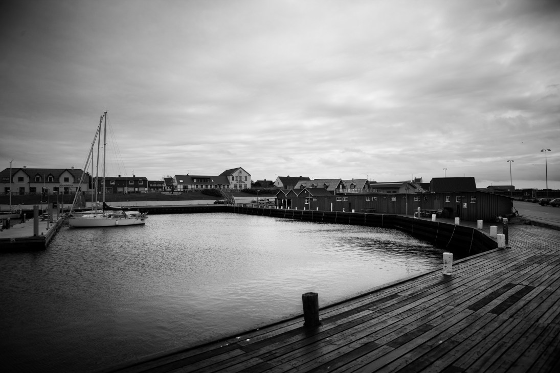 Black and white photograph of Læsø island harbor with sailboat, wooden pier, and traditional Danish buildings along the waterfront.