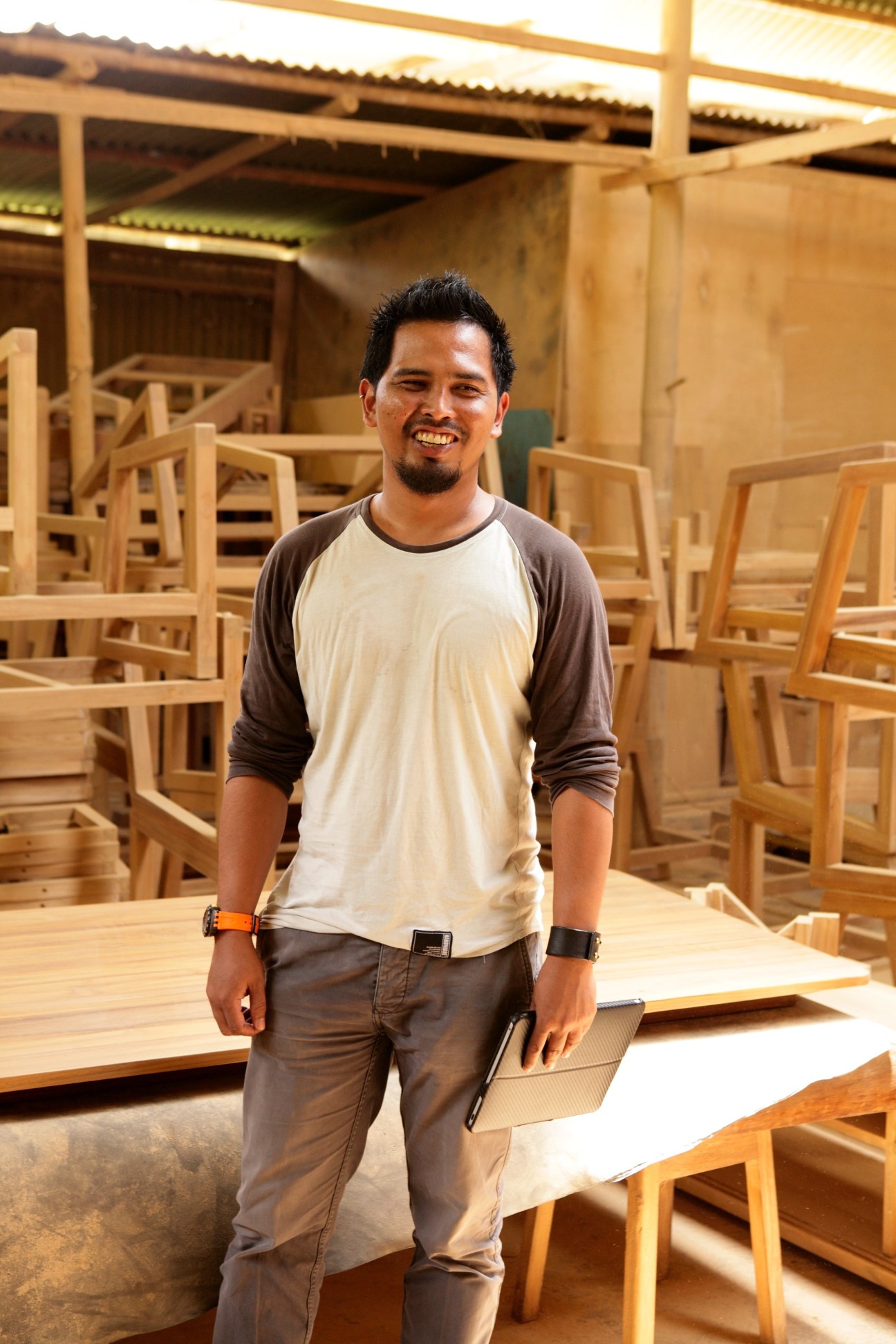 Product designer Satya Bramantya standing in his woodworking studio surrounded by wooden chairs and furniture pieces.