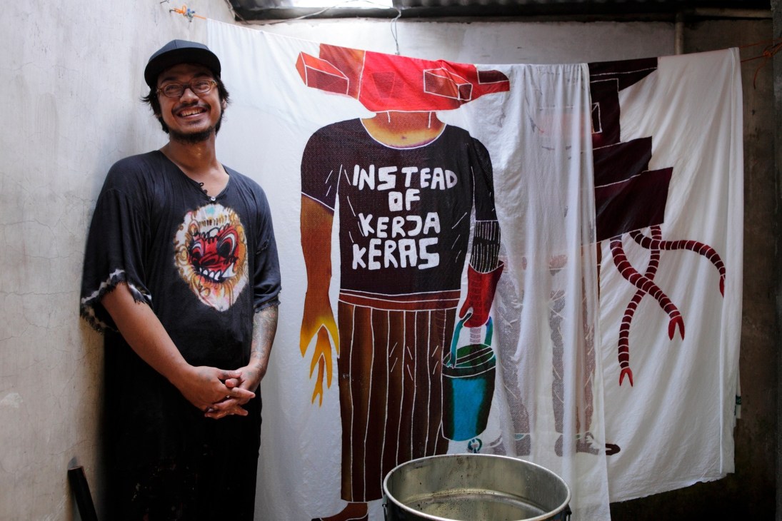 Batik-craftsman Arwin Hidayat smiling beside a colorful textile artwork with Indonesian text.