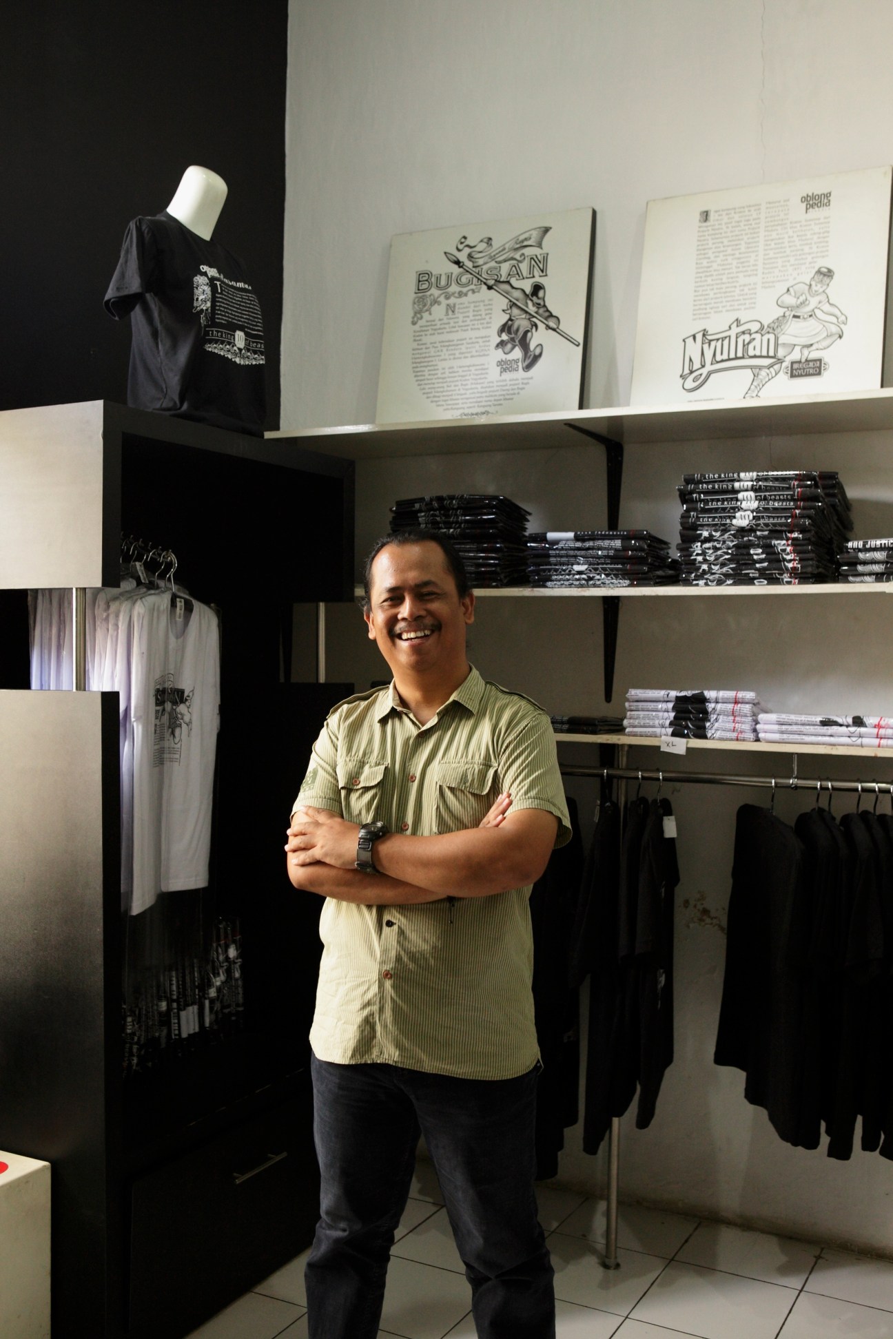 Noor Arief stands smiling in his Dagadu Djokdja factory, surrounded by shelves displaying t-shirts and merchandise.