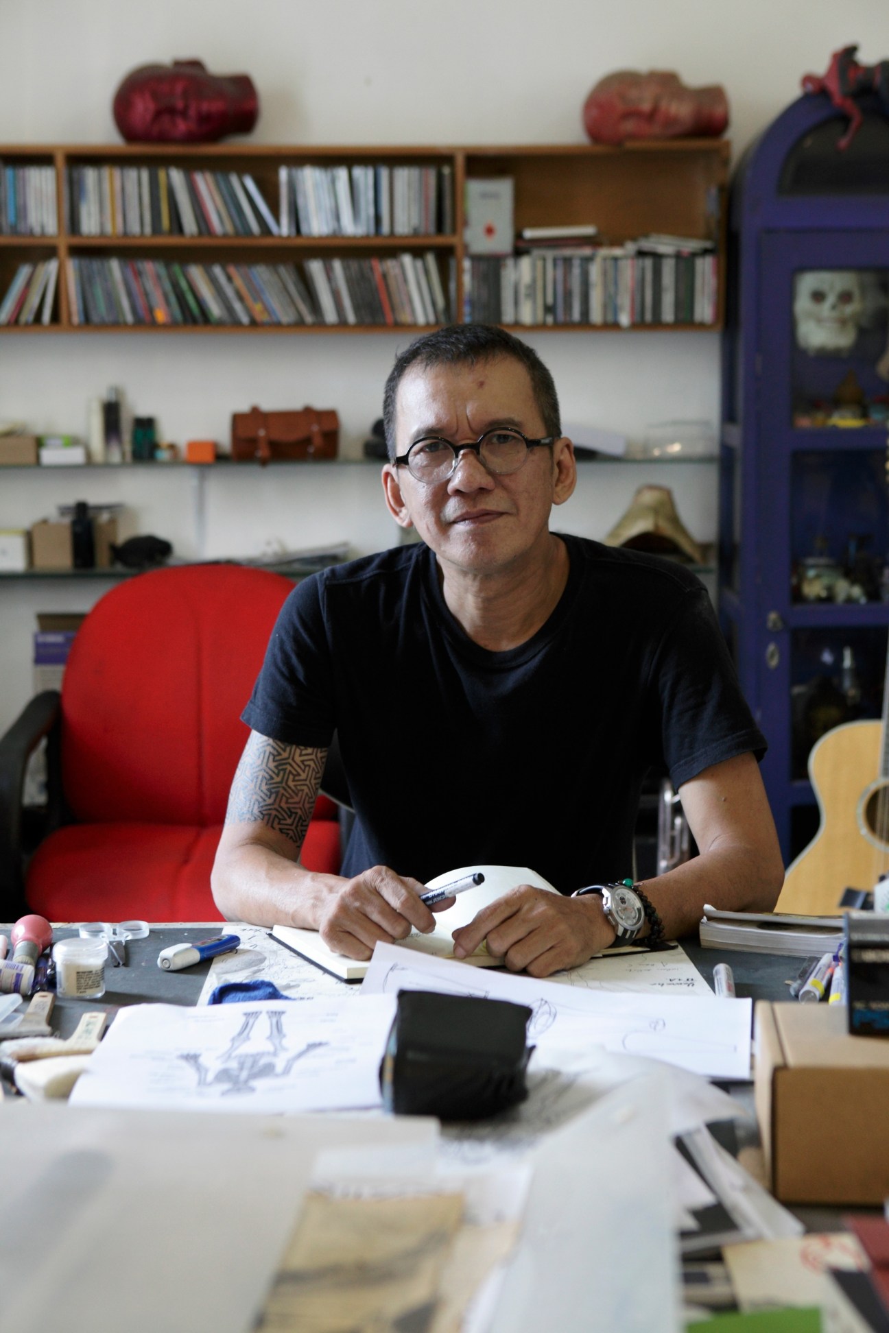 Leading contemporary artist Agus Suwage seated at his desk in his studio surrounded by shelves of CDs, books, and art materials.