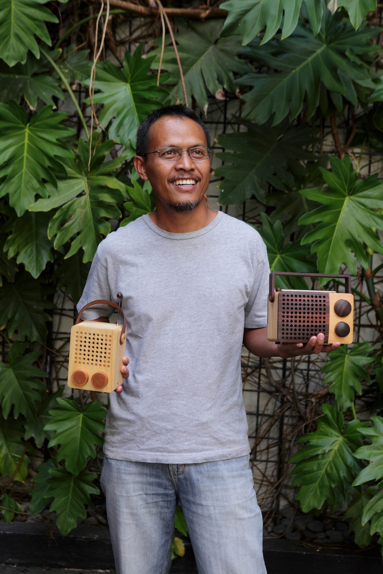 Singgih Susilo Kartono holding two wooden Magno radios in front of green foliage.