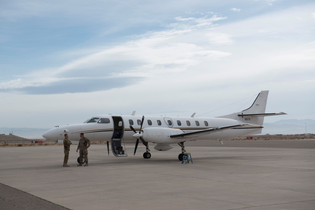 A white military turboprop aircraft parked on a runway with two uniformed personnel standing nearby.