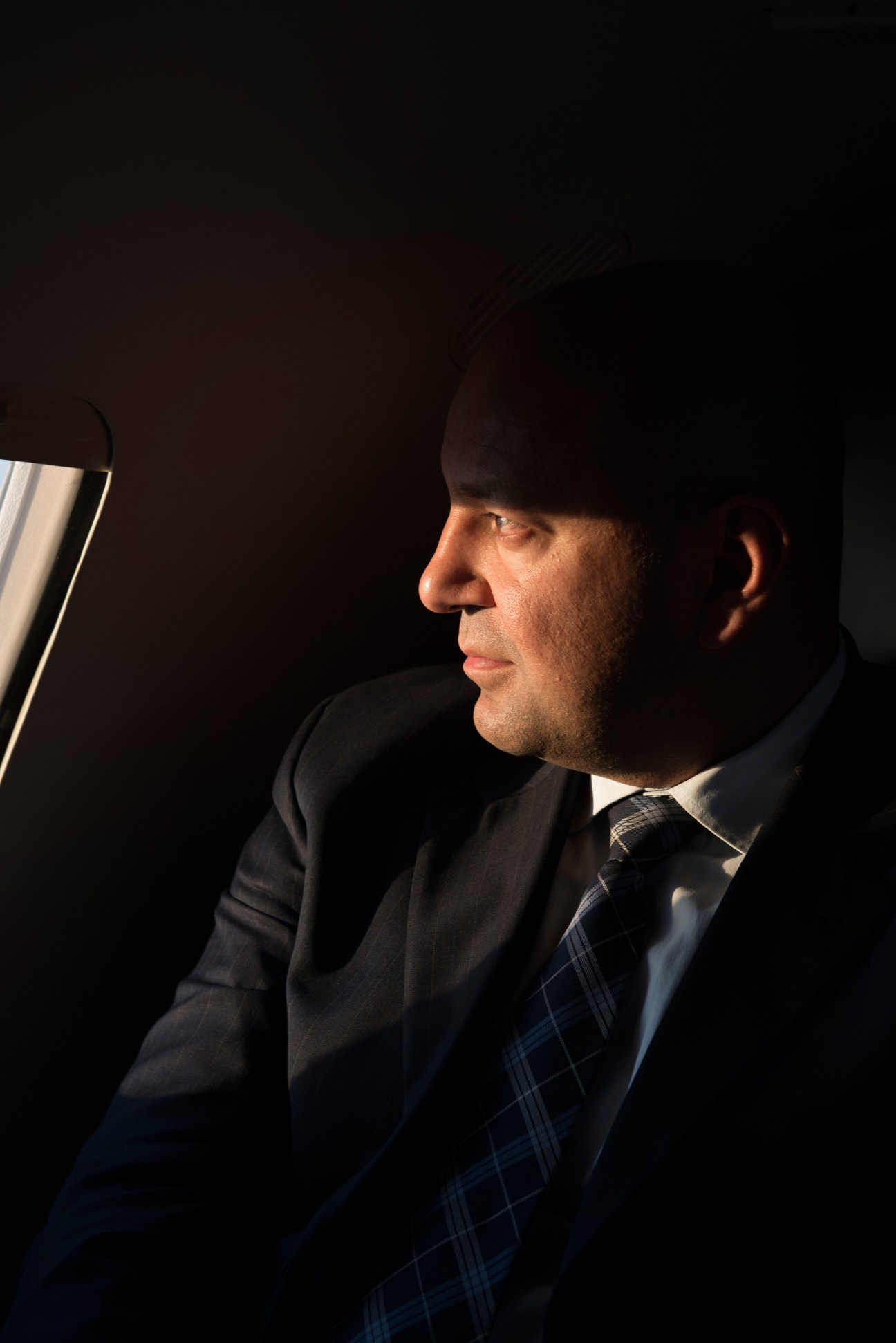 Man in dark suit and tartan tie looking to the side against dark background.