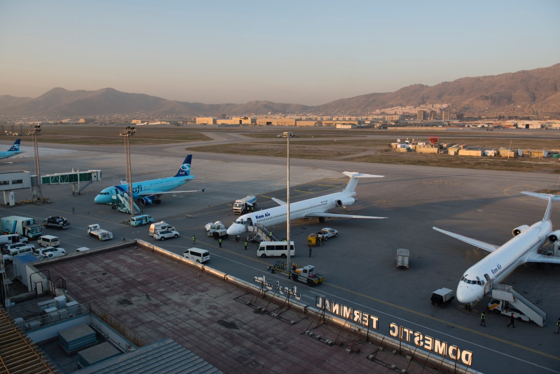 Multiple commercial aircraft parked at Kabul airport terminal with ground support vehicles and mountains visible in background.