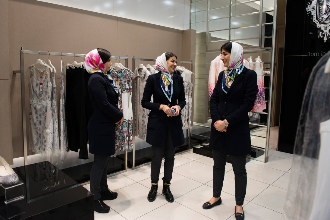 Three women in headscarves and dark coats stand in a modern clothing boutique with white garments on display racks.
