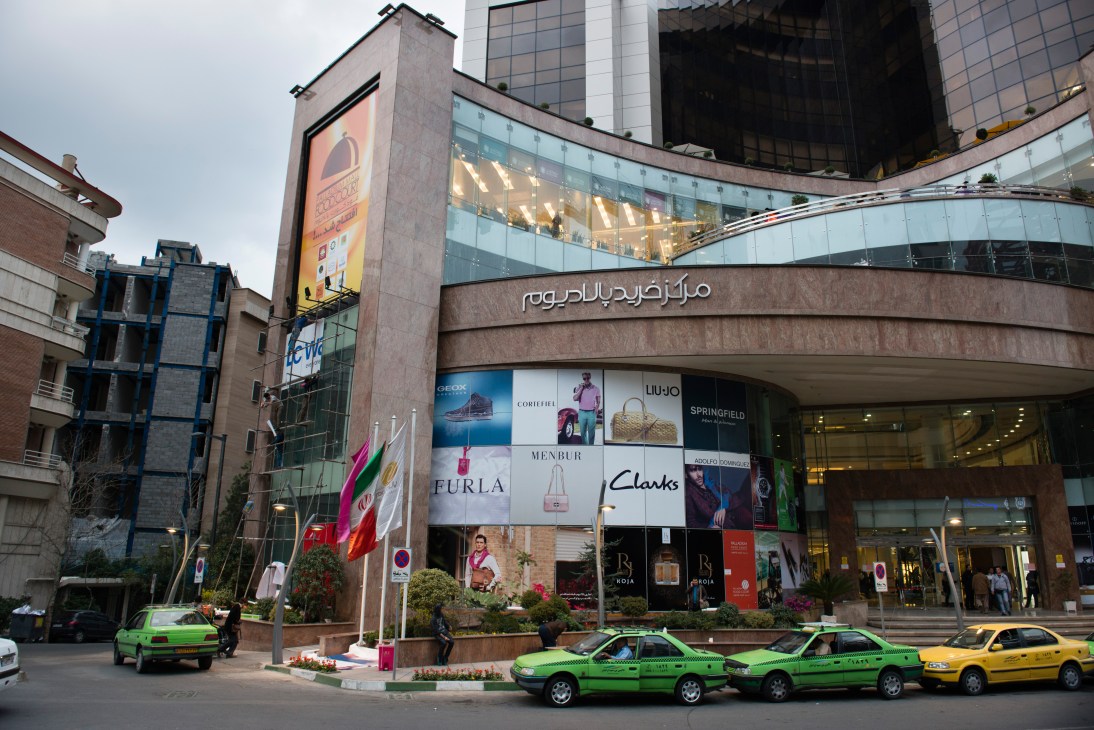 Exterior of Palladium mall in Tehran with luxury brand signage and green taxis parked outside.