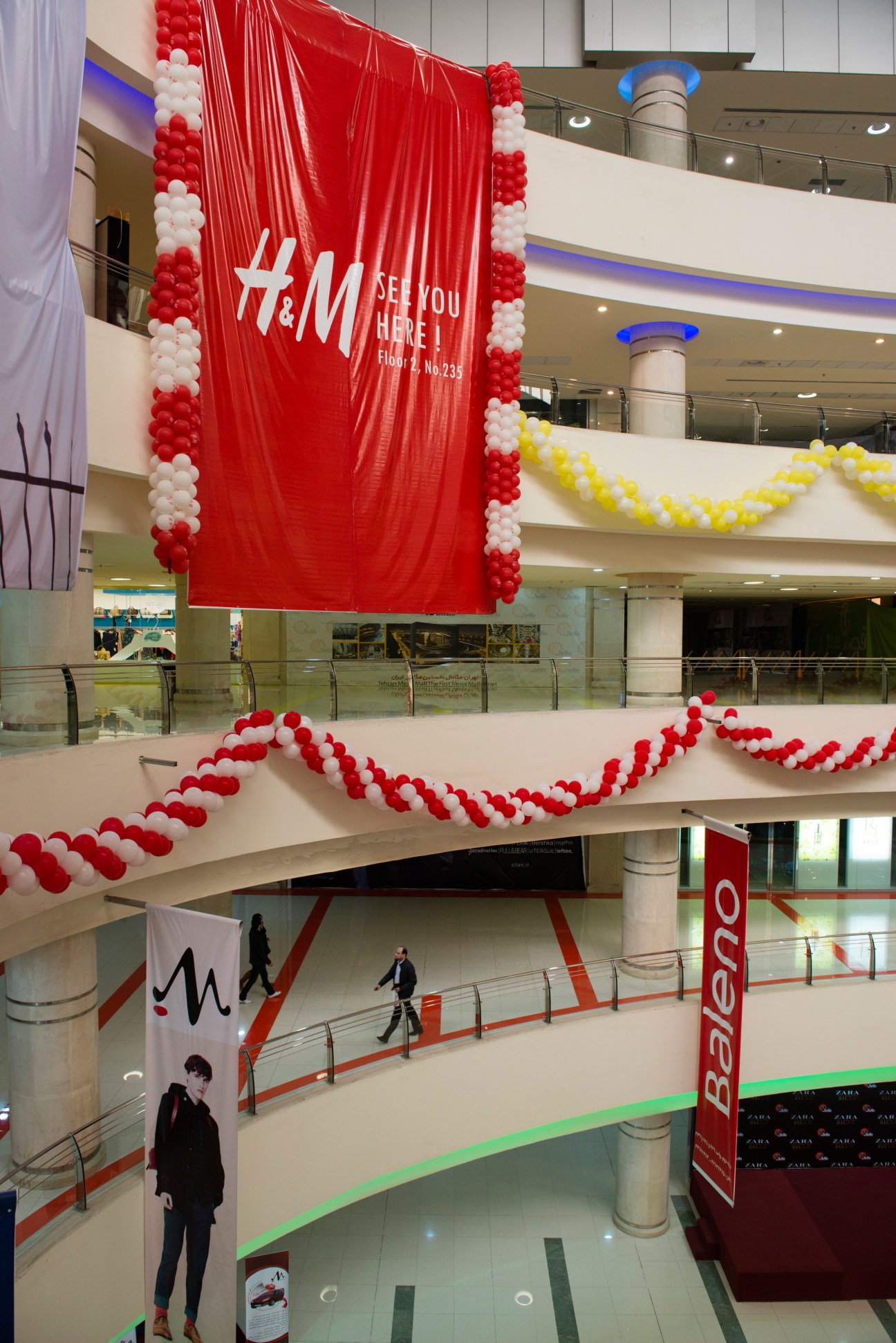 H&M storefront banner with red and white balloon decorations in a multi-level shopping mall atrium.