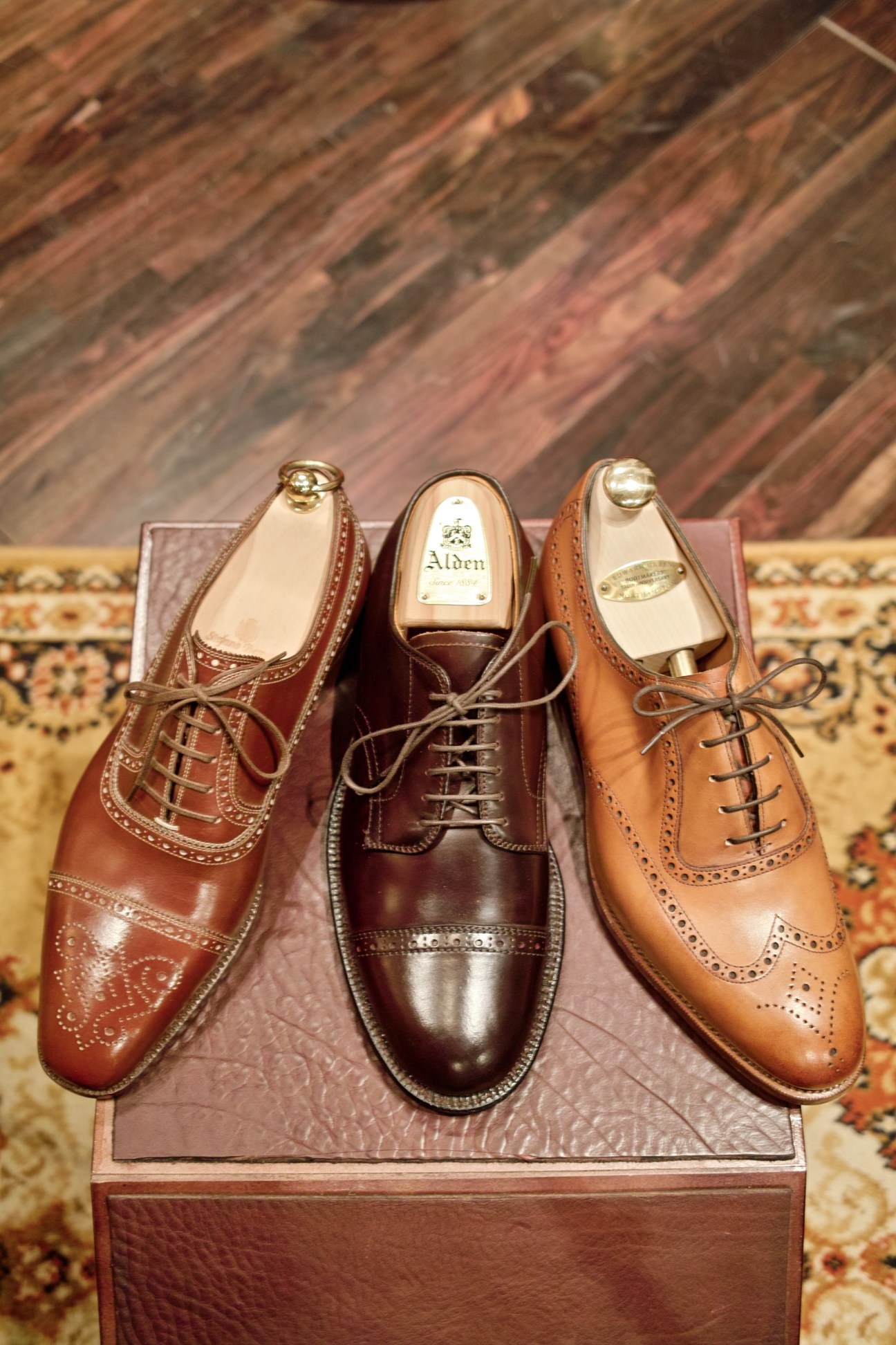 Three leather men's dress shoes in brown tones displayed on a wooden block with shoe trees.