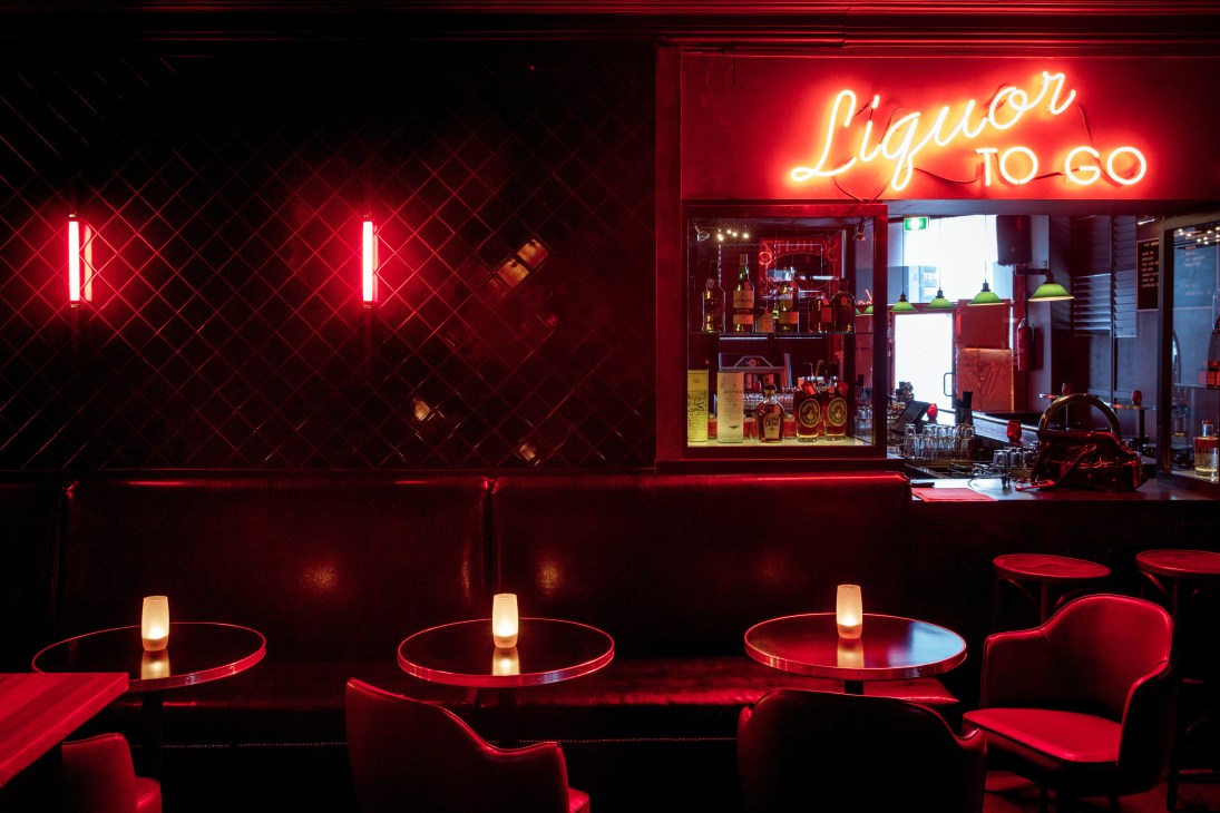 Red neon-lit bar interior with round tables, glowing cylindrical lamps, and liquor store window display