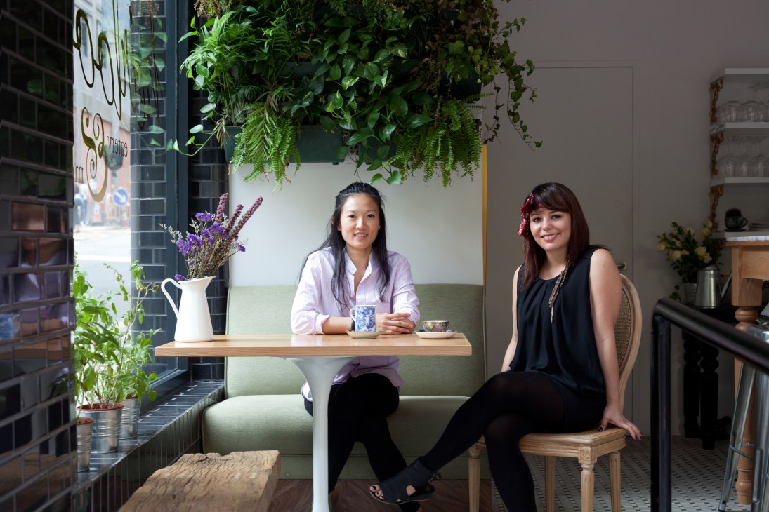 Two women in a plant-filled restaurant interior with green upholstered seating and wooden table at Heirloom in Hong Kong.