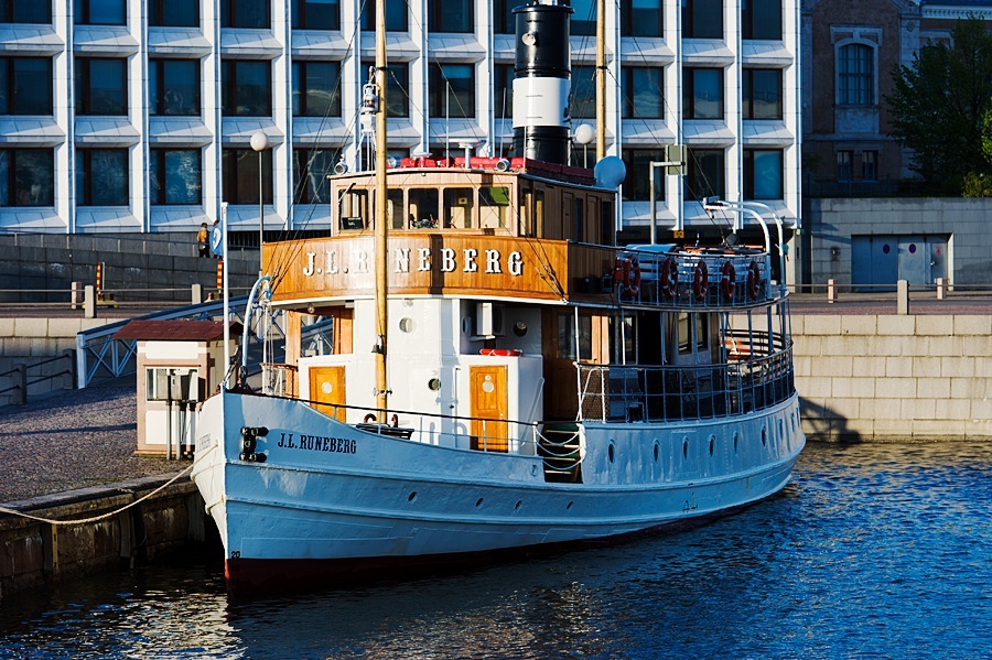 Blue and yellow ferry boat J.L. Runeberg docked at Helsinki waterfront with modern buildings behind.