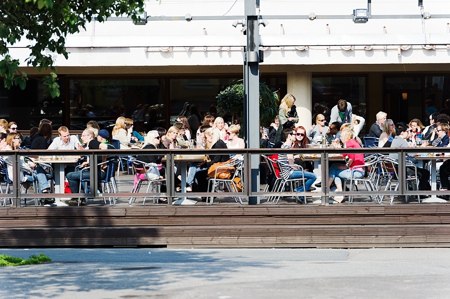 Outdoor waterfront café terrace with diners seated at tables along a wooden deck.
