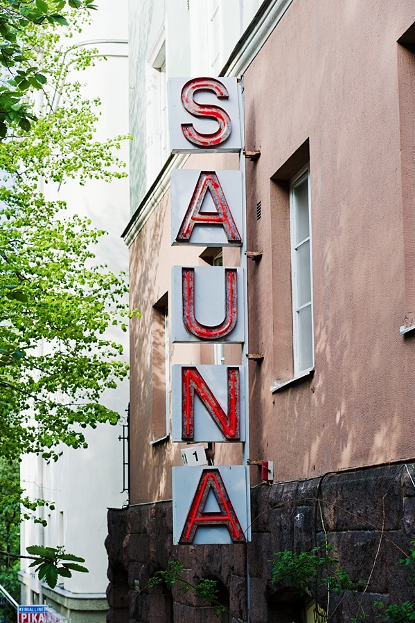 Vertical red neon sign reading 'SAUNA' mounted on a brick building with green foliage visible.