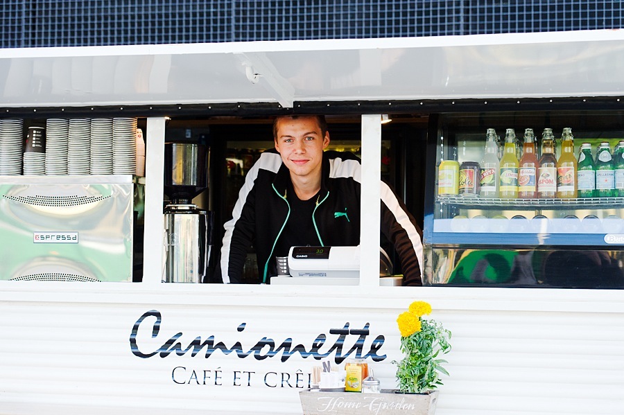 Young barista stands at the counter of a white mobile café called Camionette with beverage coolers and espresso equipment visible behind him.