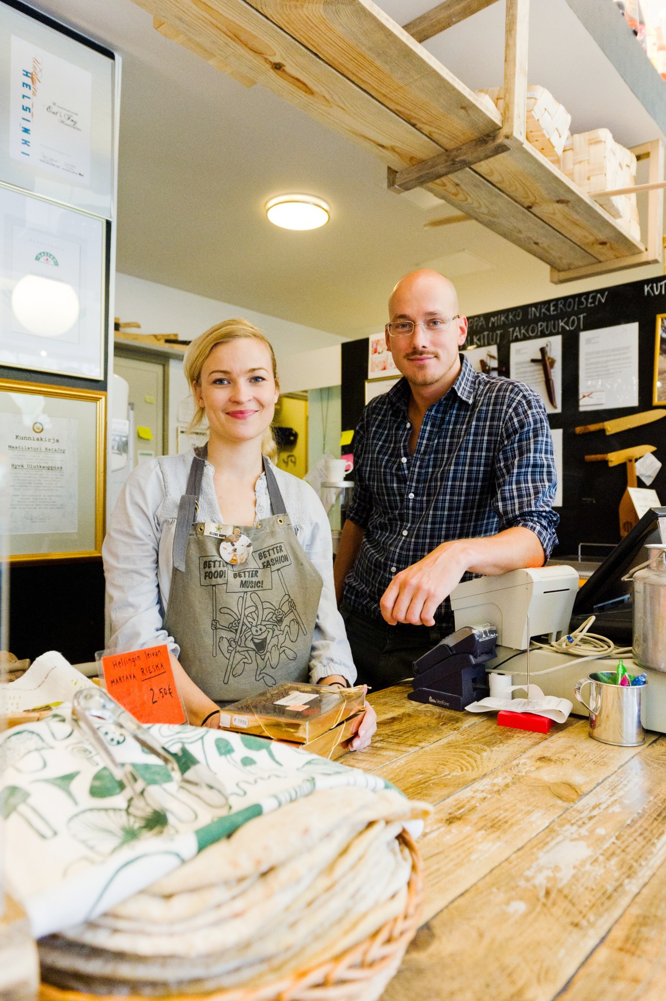 Two people standing behind a wooden counter in a small shop with exposed wooden beams and shelving.