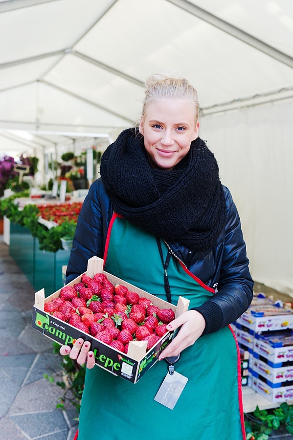 Woman holding a crate of fresh raspberries in a greenhouse market setting.