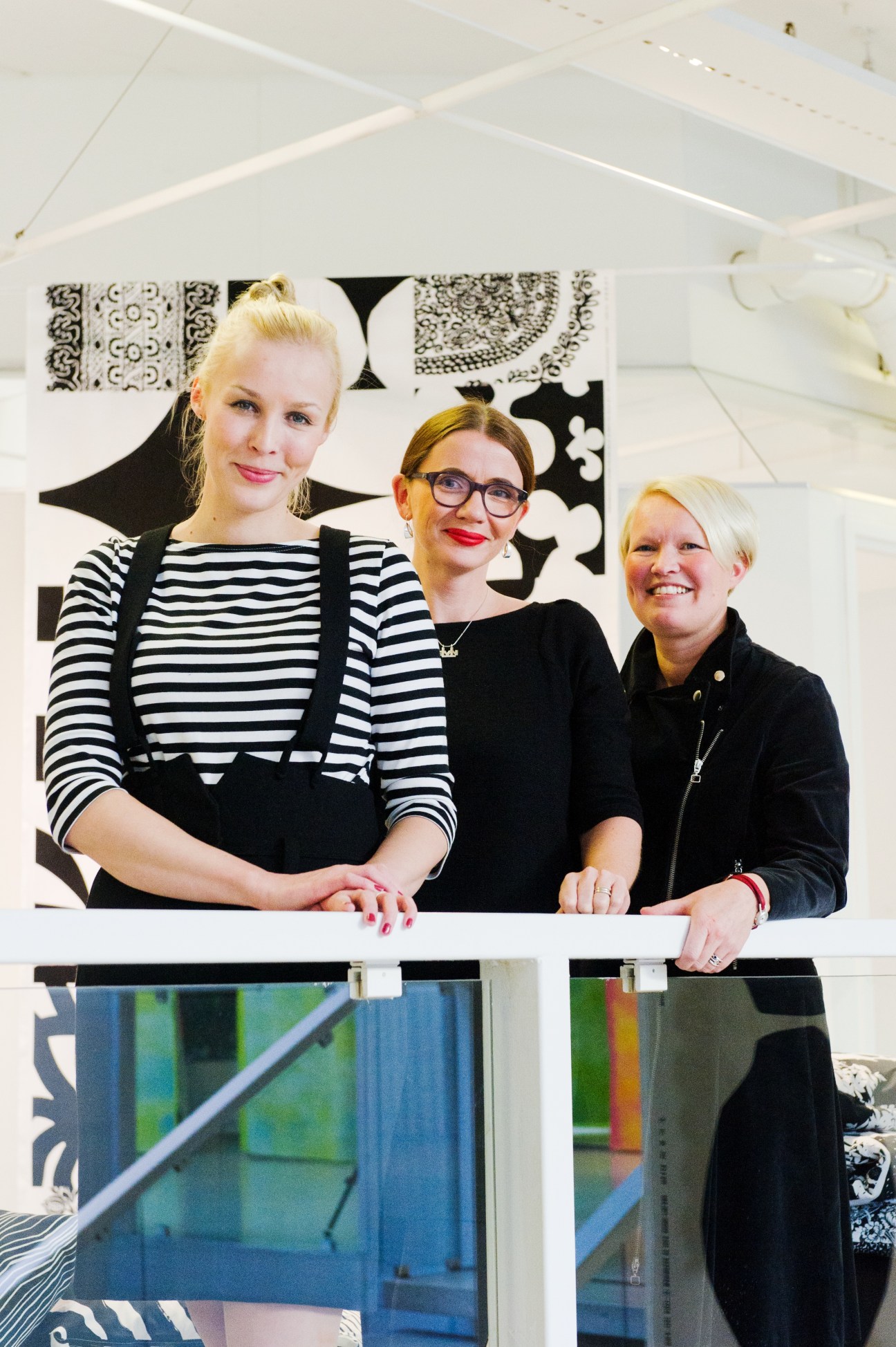 Three women posing in front of a white wall with black and white patterned design elements and colorful panels below.