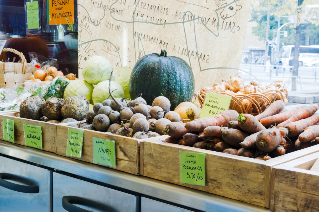 Helsinki market stall displaying fresh vegetables including carrots, beets, cabbage and melon with price labels in Finnish.