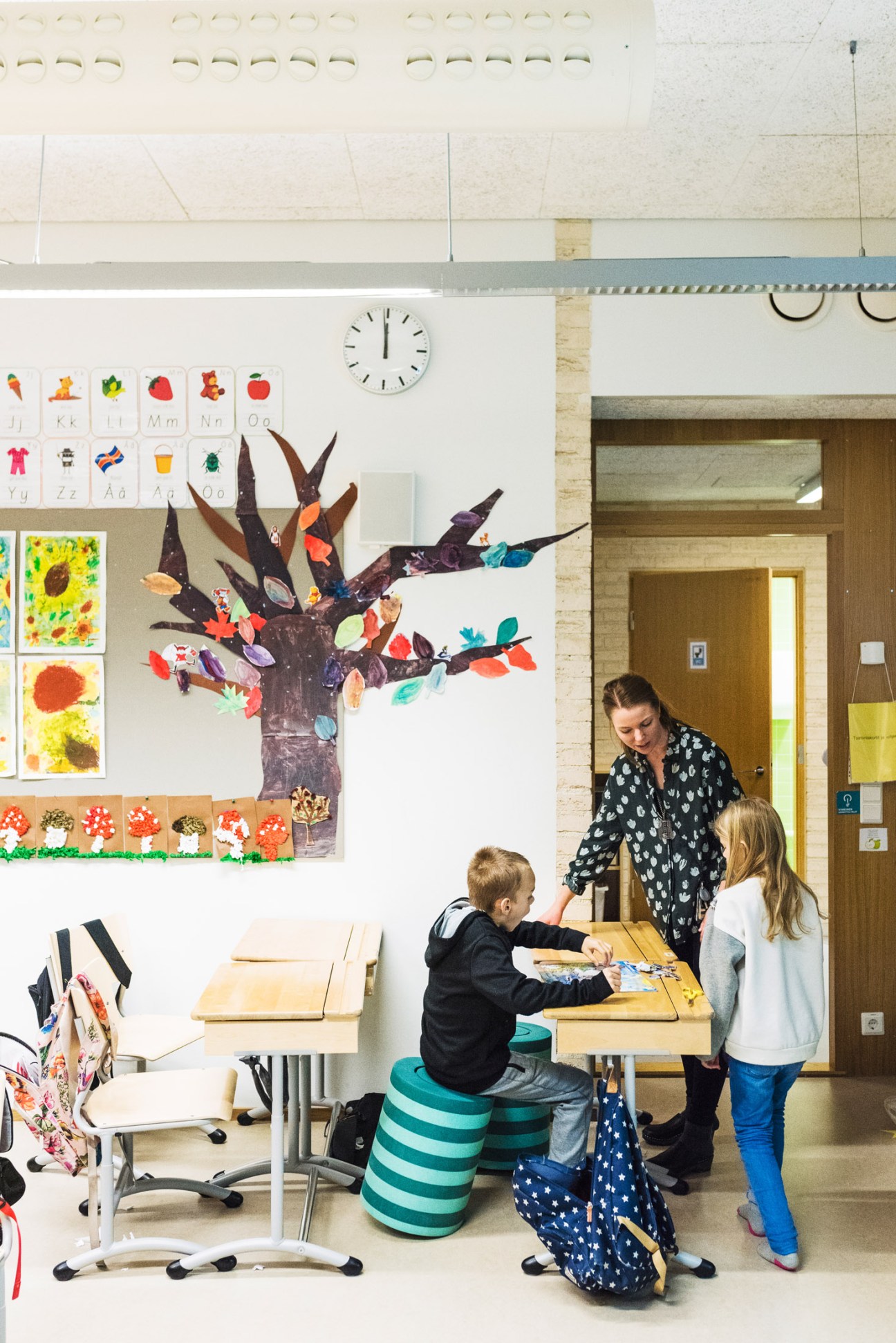 Teacher guides two children at a wooden desk in a bright classroom decorated with student artwork and a colorful tree mural.