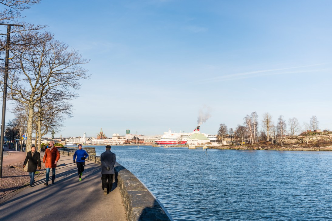 People jogging and walking along a waterfront promenade in Helsinki with ships and buildings visible across the bay.