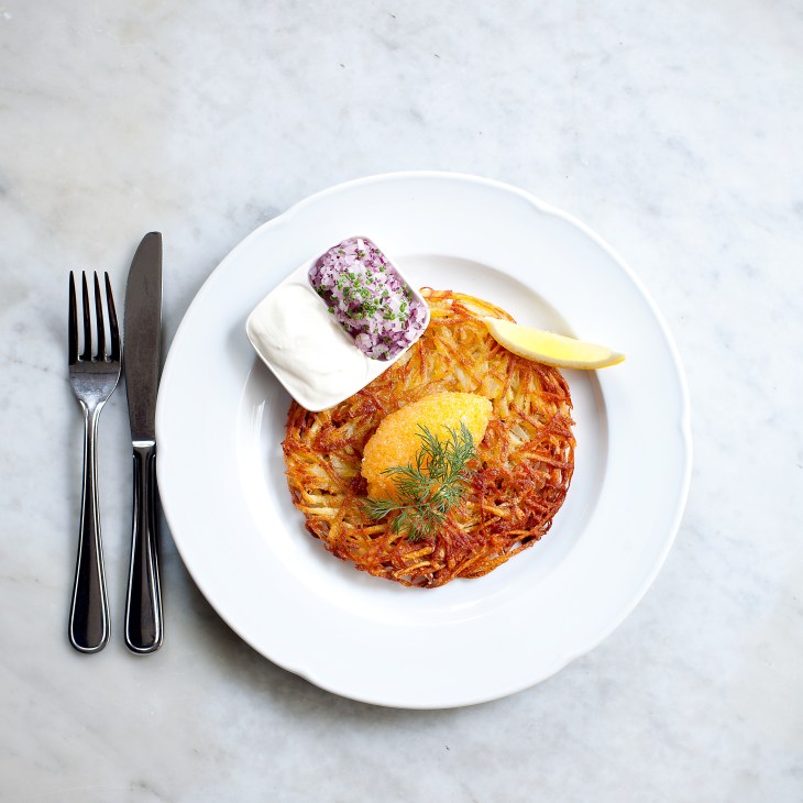 Råraka potato rosti topped with Kalix Löjrom roe, sour cream, red onion and dill on white plate.
