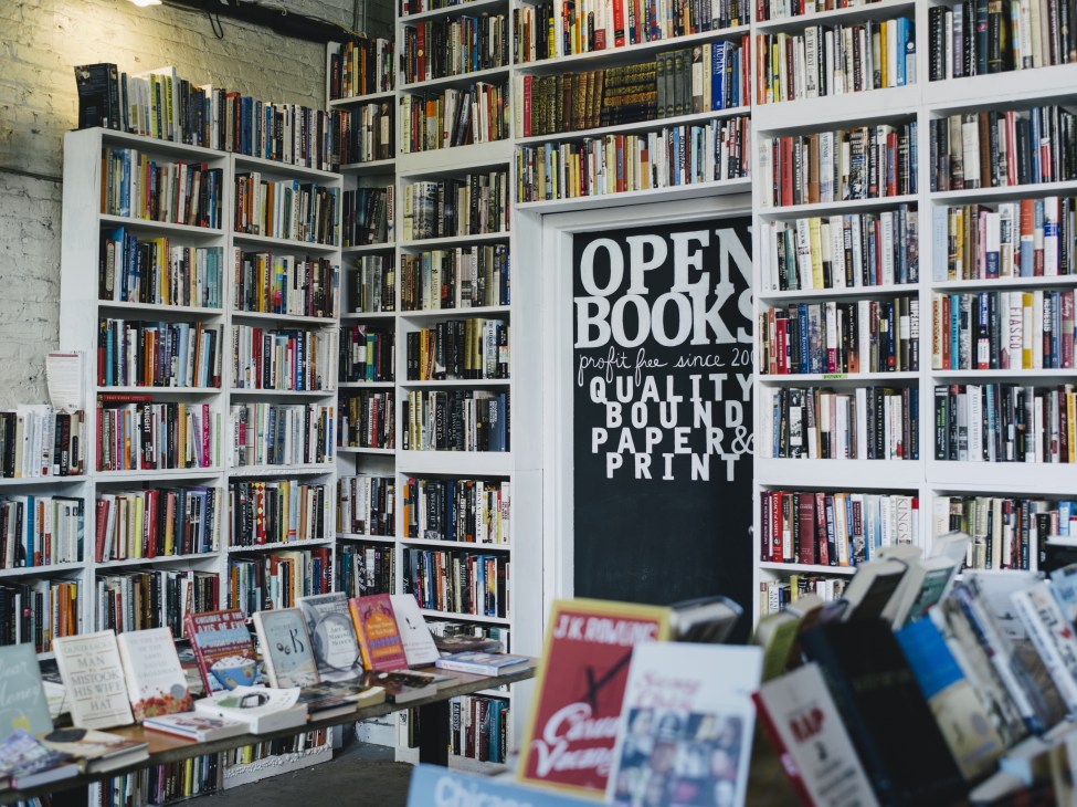 Interior of Open Books used bookstore showing white shelving units stocked with books and a black sign reading 'OPEN BOOKS'.