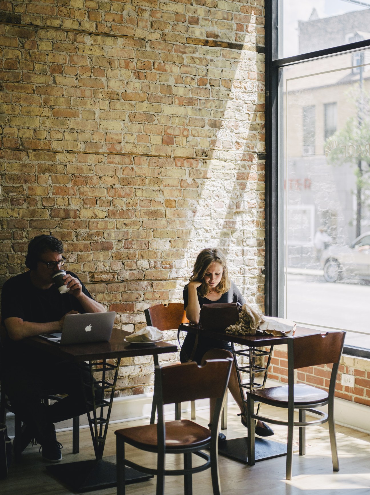 Two people work at tables inside Beurrage bakery with exposed brick walls and large windows.