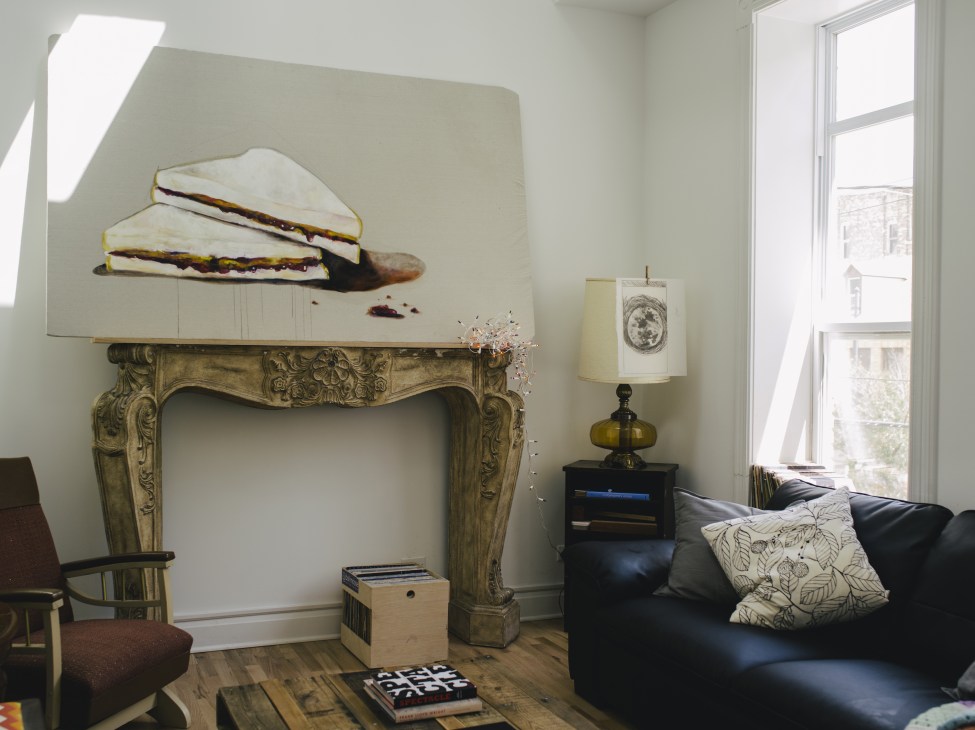 Modern living room with ornate fireplace mantel, black sofa, and sandwich artwork on white canvas.