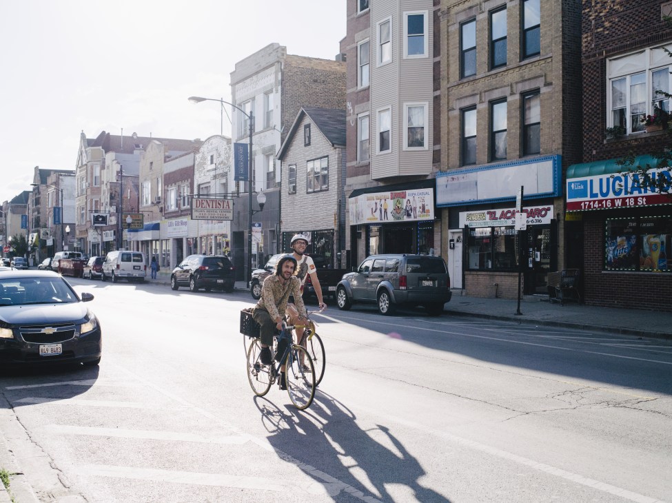 Two people riding a bicycle down 18th Street in Chicago's Pilsen neighborhood with storefronts and parked cars visible.