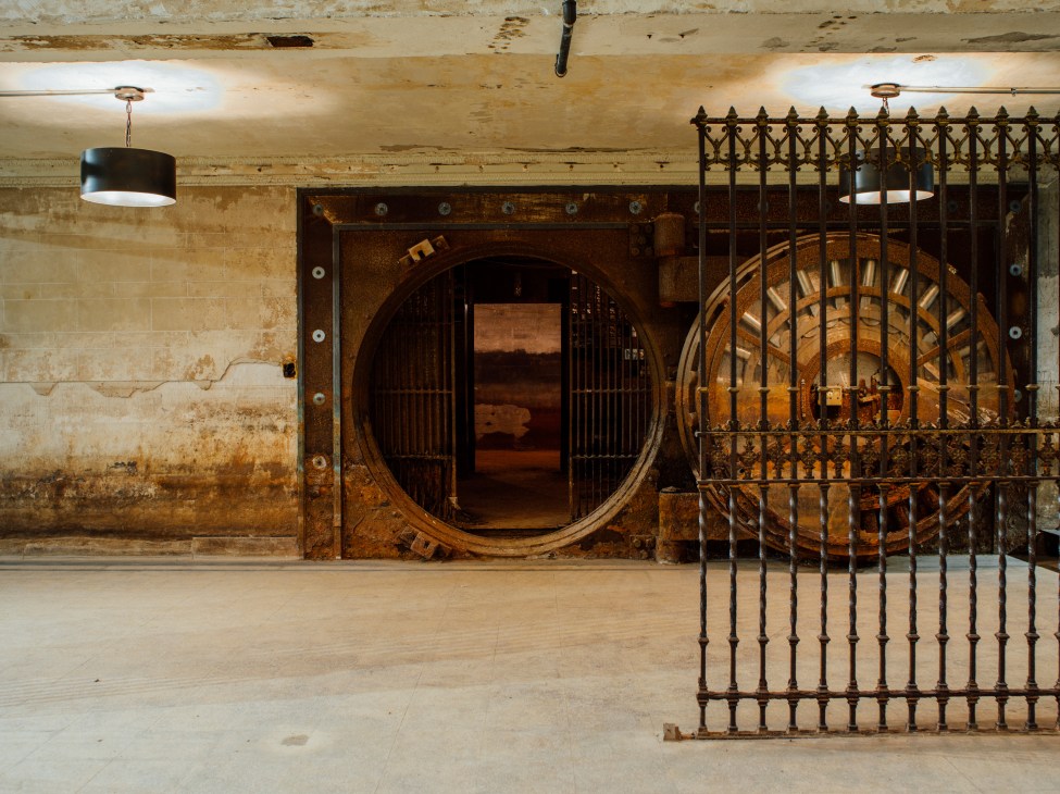 Large circular rusted bank vault door in basement with ornate iron gate and pendant lighting.