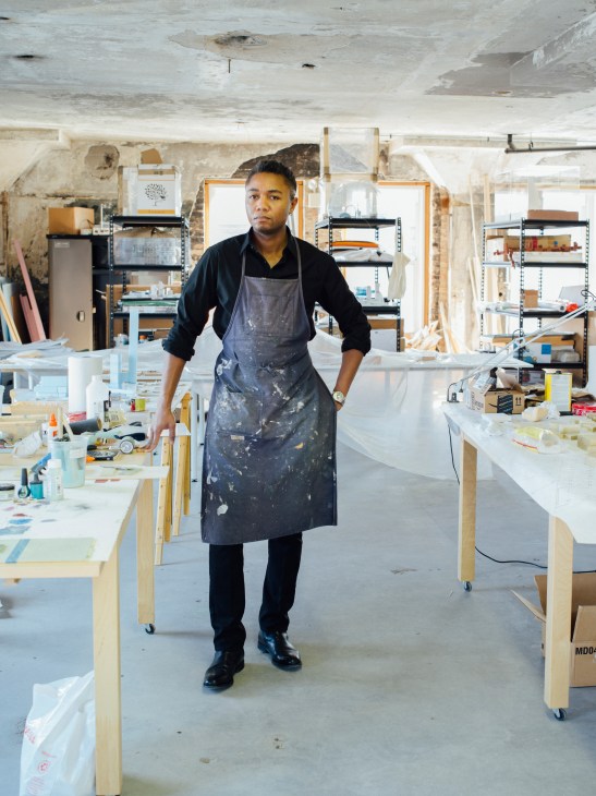 Architect Marshall Brown stands in his studio wearing a work apron, surrounded by wooden work tables and shelving.