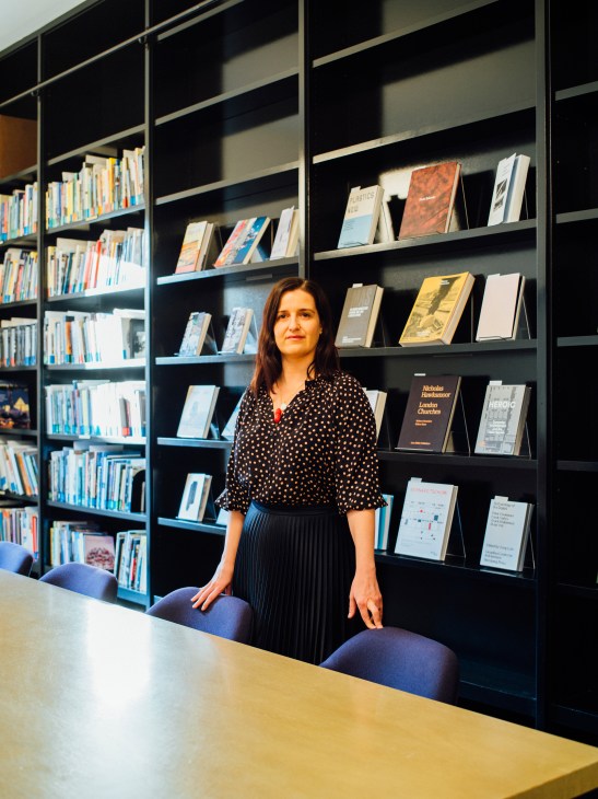 Sarah Herda of the Graham Foundation stands before a wall of shelves filled with architecture and design books.