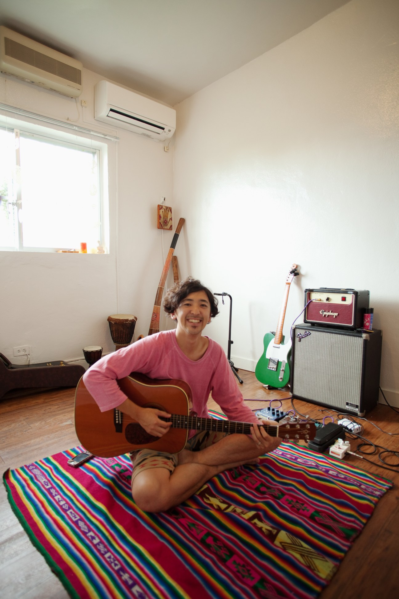 Yusuke Hashizume sits smiling with an acoustic guitar in a spacious room with musical instruments and amplifiers.