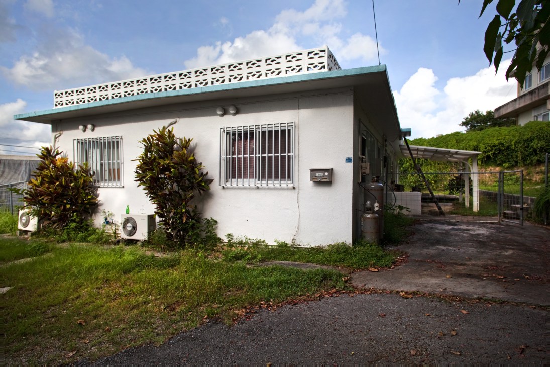 Single-story concrete bungalow with turquoise trim, barred windows, and surrounding vegetation in Okinawa.