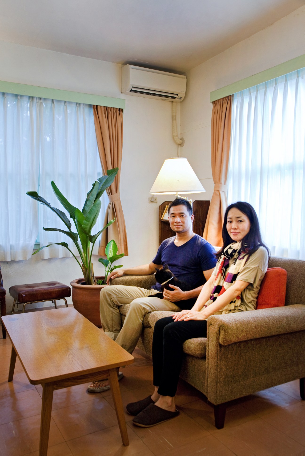 Toshikatsu and Azusa Saji sit on a sofa in their living room with large windows and air conditioning unit visible.
