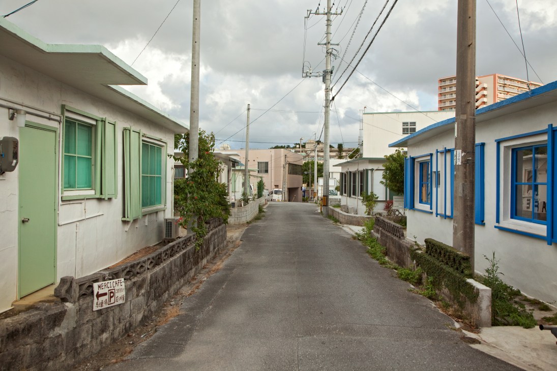 A quiet street lined with single-story concrete military bungalows painted in pastel green and blue in Okinawa.