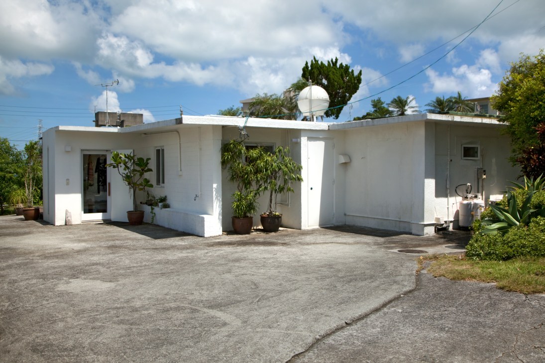 White concrete single-story bungalow with flat roof, large windows, and potted plants in front yard.