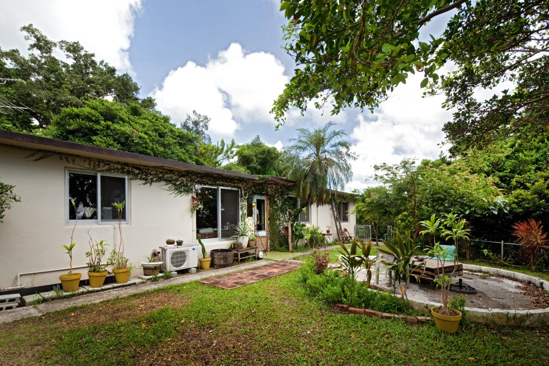 White concrete single-story bungalow with large windows, surrounded by lush green garden and tropical plants.