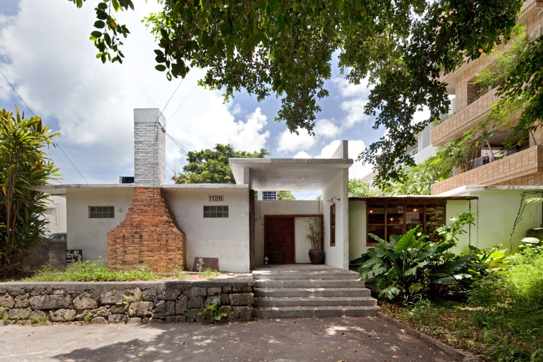 A single-storey concrete bungalow with a brick chimney, numbered 1126, featuring a covered entrance and lush tropical gardens.