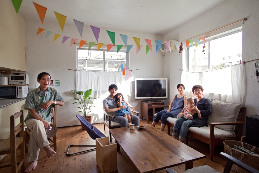 Go Maejima and family members relax in a bright living room with large windows and colorful bunting decorations.