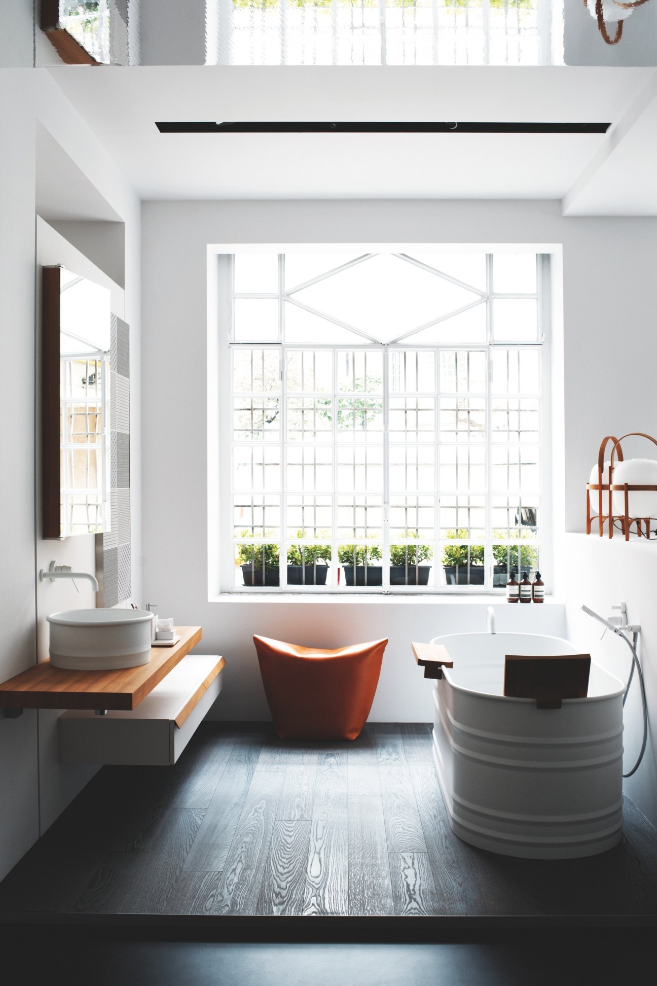 Modern bathroom showroom with white circular bathtub, red chair, wooden sink counter and large window with plants.