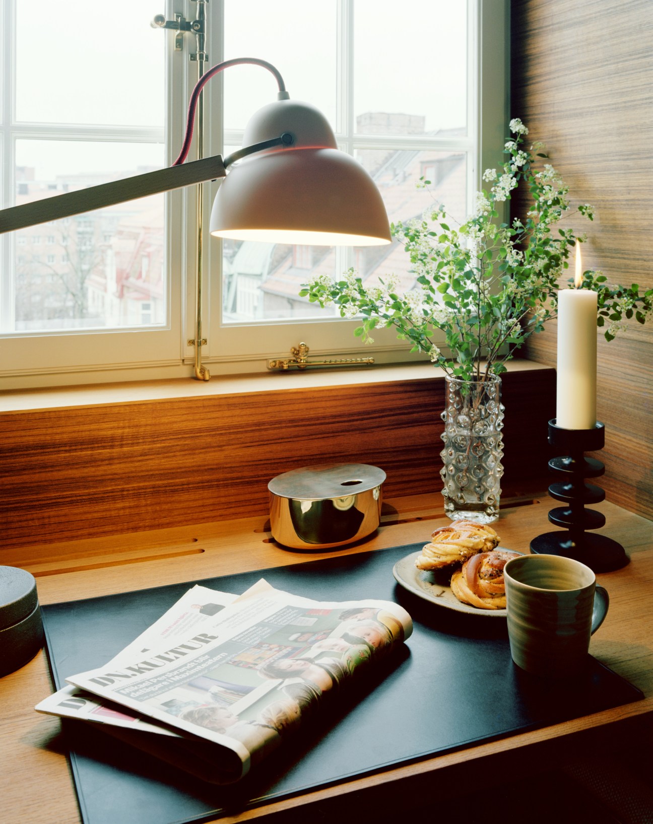 A hotel room windowsill with a brass arc lamp, fresh flowers, candle, coffee cup, pastries and newspaper in natural light.