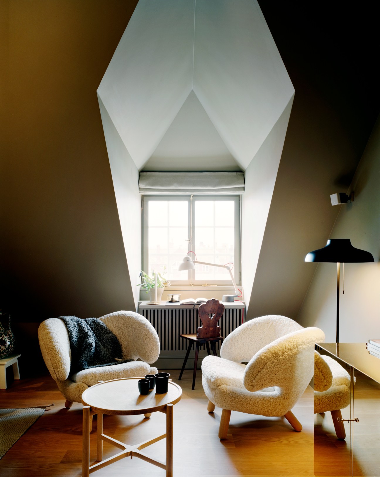 A bright attic bedroom with sloped white walls, featuring two cream-colored armchairs, a small round table, and a reading nook by a skylight window.